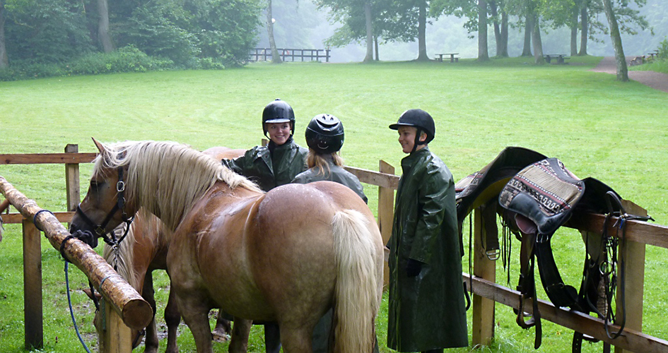 Två hästar och tre tjejer med ridhjälm och regnkläder står vid hästuppställningsplatsen i Skäralid. Bakom inhägnaden finns en grön gräsmatta som omsluts av skog.